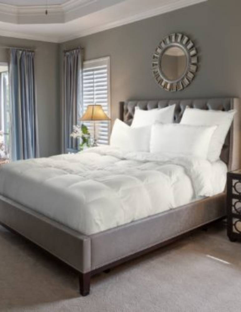 A neatly made bed with a plush white comforter and multiple white pillows is the main focus. The bed has a tufted headboard and sits in a softly lit bedroom with gray walls, blue curtains, a round decorative mirror, and a bedside lamp on a nightstand. The room feels calm and inviting, with a peaceful and comfortable atmosphere. No visible text is present in the image.